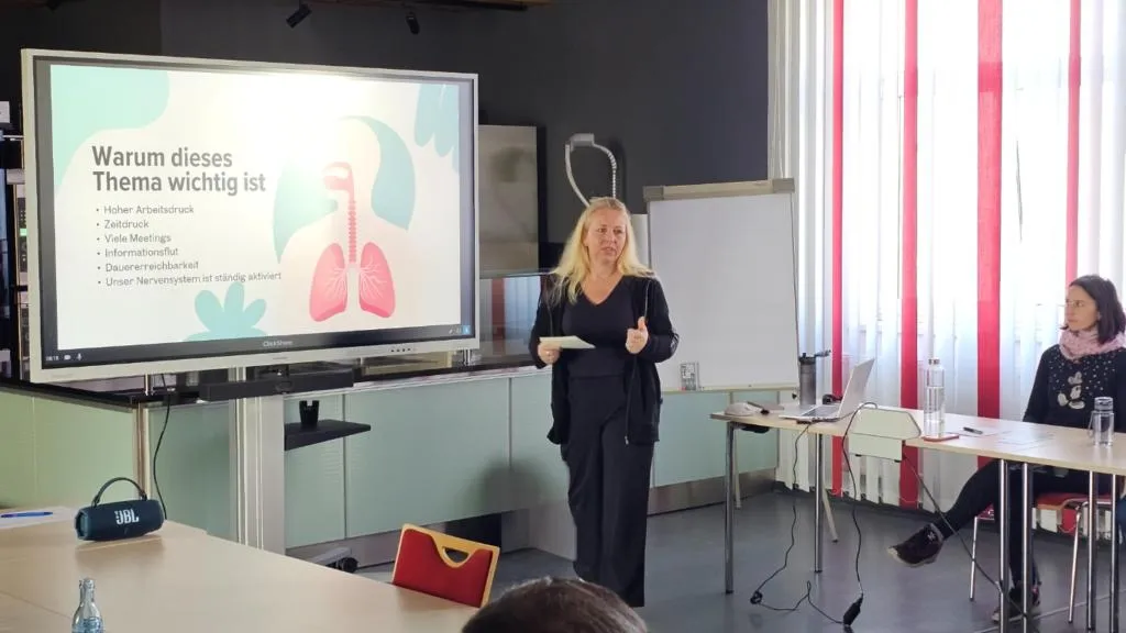 Ines Richter hält einen Atemworkshop vor Mitarbeitenden, auf dem Bildschirm eine Folie über Arbeitsdruck und das Nervensystem